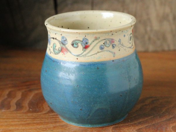 Bauchiger Becher mit aufgemalter Blumenranke auf hellem Untergrund im oberen Drittel, unten petrol glasiert, innen transparent glänzend.