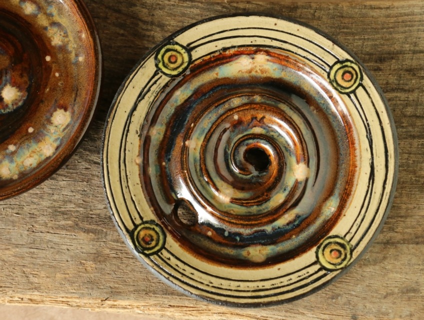 Seifenschale mit 4 bunten Kreisen am hellen Rand, die durch Linien verbunden sind. Die Mitte, wo die Seife aufliegt ist rotbraun mit hellen Effekten glasiert.